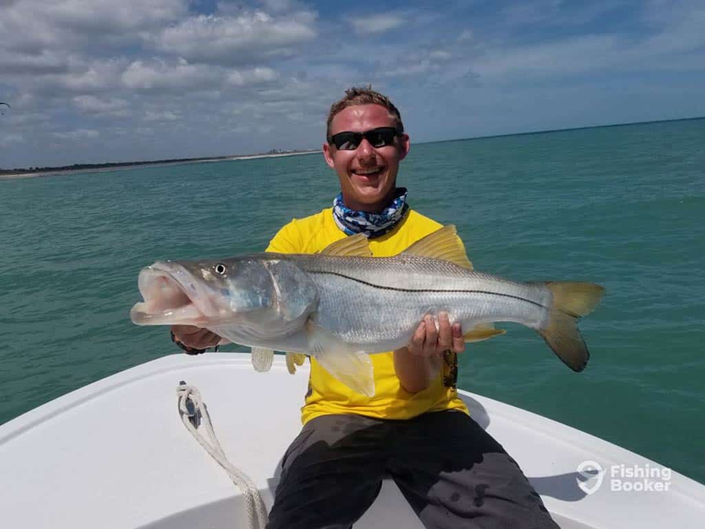 An angler wearing a yellow shirt sits at the front of a fishing boat, holding a large Snook caught in the inshore waters near Vero Beach on a sunny day, with the clear waters behind him