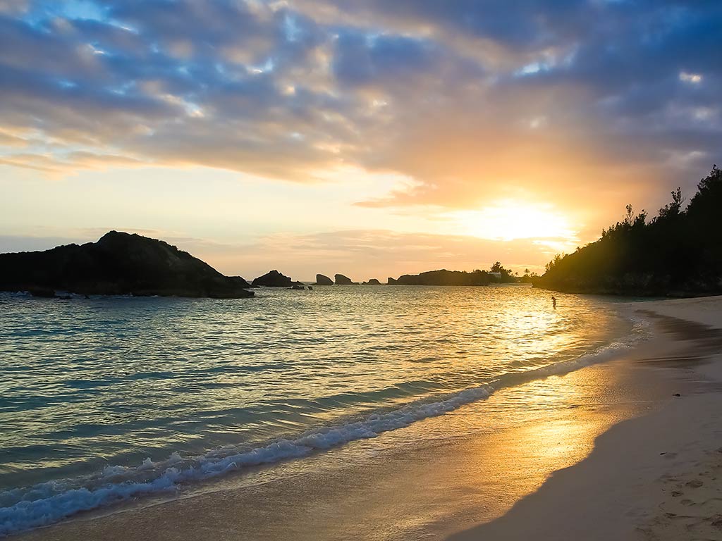 A view across a beach in Bermuda at sunset, with the calm waters on the left of the image and the sands curving round to some rocks on the right