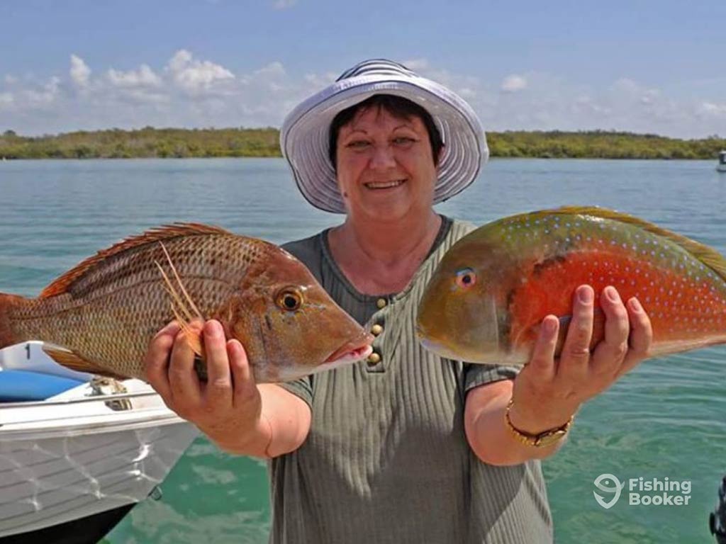 A female angler in a summer had, holding up a Sweetlip Emperor and another small fish on a clear day in the backcountry waters of Moreton Bay