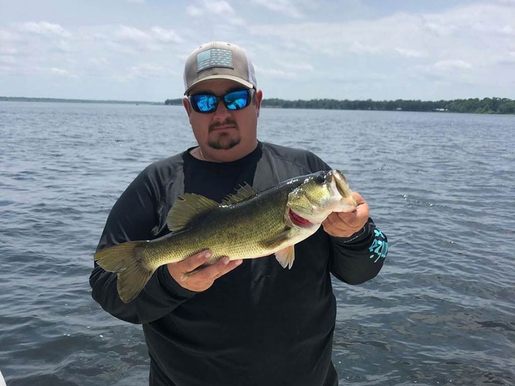 An angler in a baseball cap and sunglasses , standing in front of a river near Crystal River, holding a Largemouth Bass on a sunny day