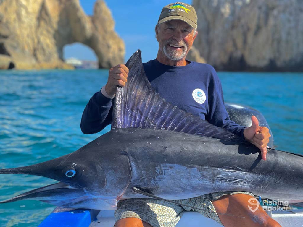 A closeup of an elderly angler in a blue sweater and green baseball cap holding a large Marlin while sitting on a fishing charter in front of Los Arcos, Cabo San Lucas, on a clear day