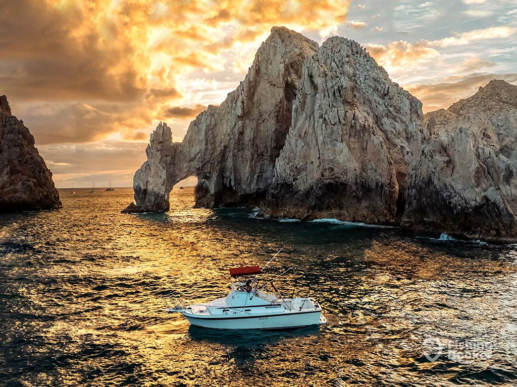 An aerial view of a fishing charter in front of Los Arcos, Cabo San Lucas, at sunset on a cloudy day with the sun setting behind clouds in the distance behind the arch