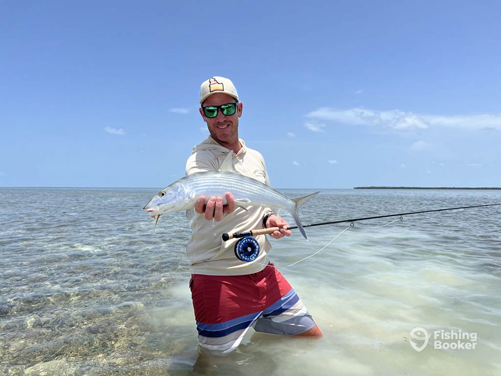 A man standing just over knee-high in the clear, sandy waters of the Florida Keys, presenting a Bonefish to the camera in one hand and holding a fly fishing rod in the other on a clear day