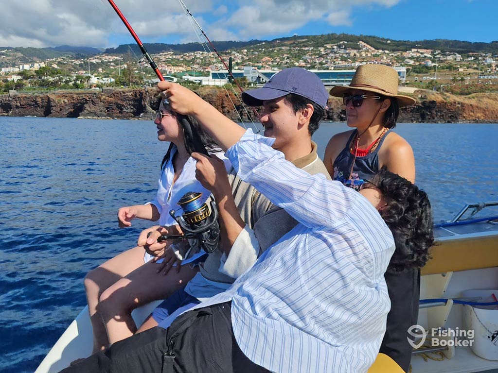 A woman angler struggling with a fishing rod off the back of a fishing charter, while others sit next to her, enjoying the spectacle on a sunny day with Madeira's land visible in the distance