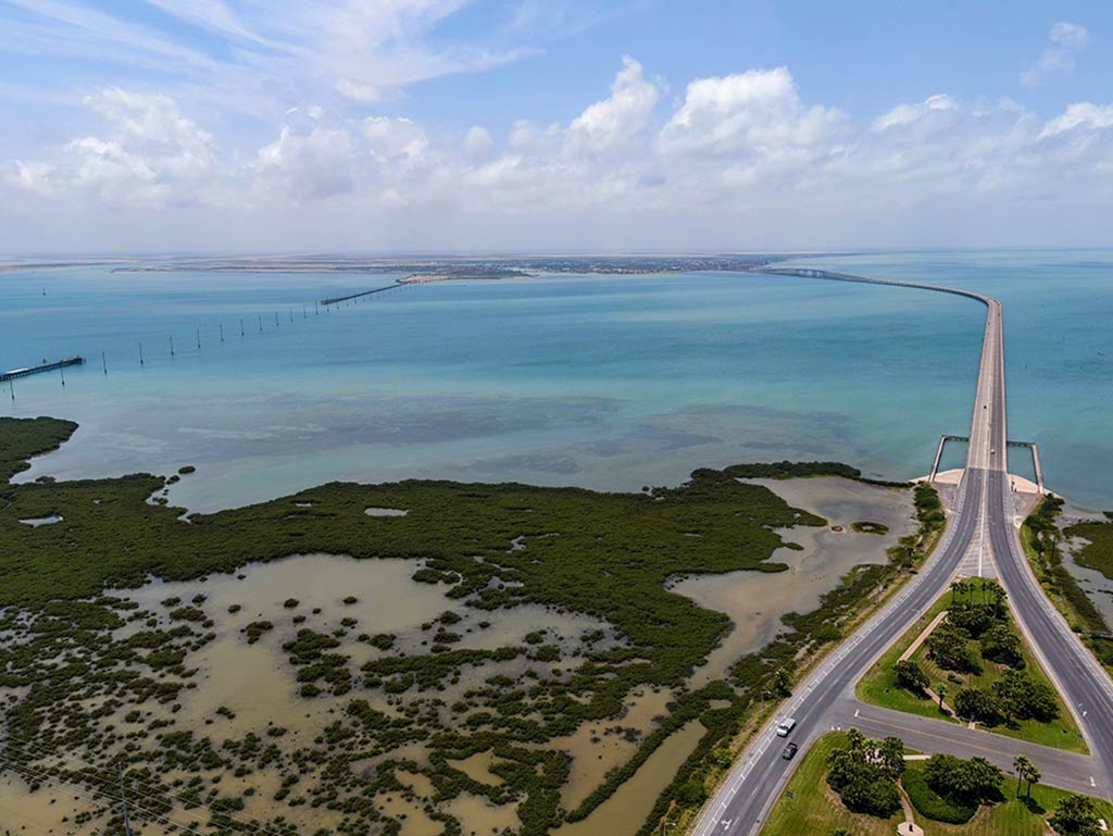 An aerial view looking towards a bridge over the Laguna Madre in Texas on a cloudy day, with murky waters visible in the foreground