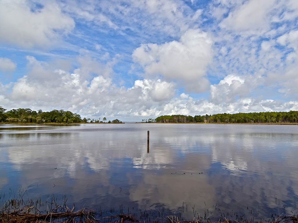 A view across the calm waters of the Choctawhatchee Bay on a day with sunny intervals, with tree-lined shores visible on either side and a pole sticking out of the middle of the water