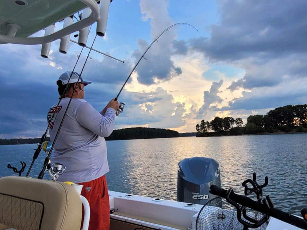 An angler casting his line over the side of a boat in Lake Lanier just after sunset, with clouds visible above the shoreline in the distance and the calm lake visible in the middle of the image