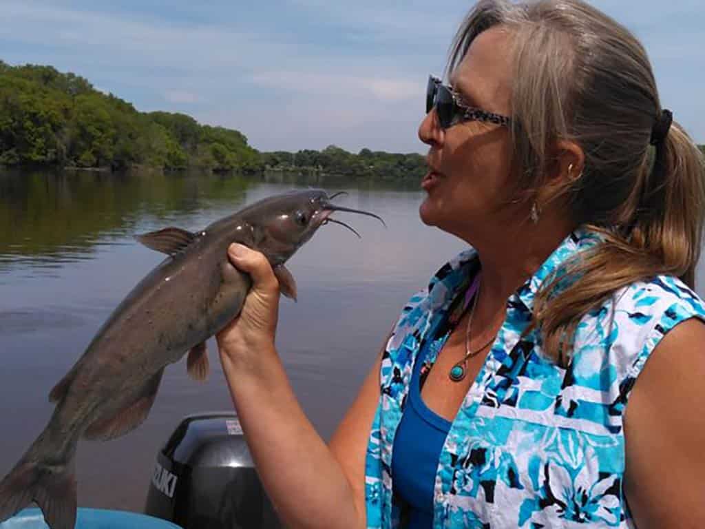 A woman in sunglasses holds up a small Catfish and pretends to kiss it on a sunny day, with water and a tree-lined shore behind her