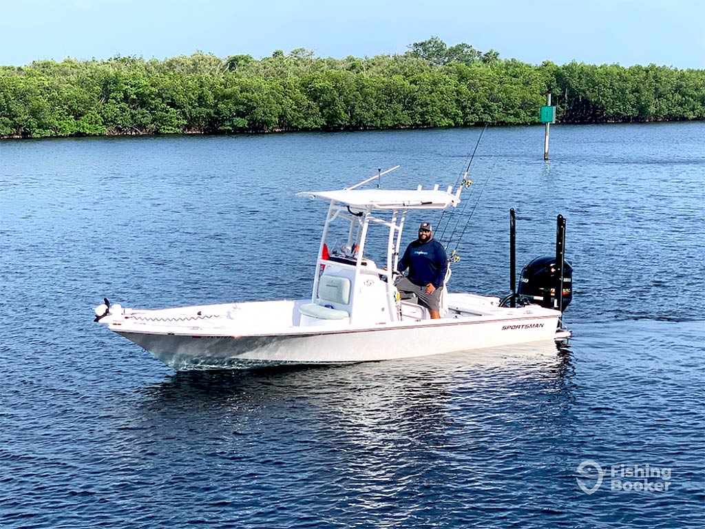 A captain steers his center console fishing boat along the inshore flats in Florida on a clear day, with a tree-lined shore visible behind him