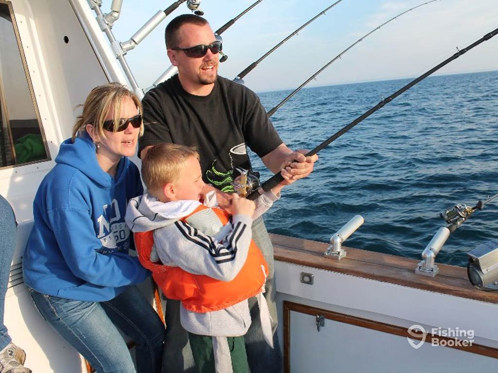 A mother and father help a little boy in a life vest with a large fishing rod in Illinois on a clear day, with the water visible behind them