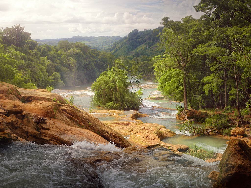 A view looking down a rocky stream in Chiapas, Mexico, with wild, lush, green foliage around fast-running water on a hazy day