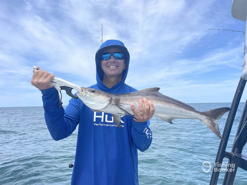 An angler in a blue hoodie and sunglasses, standing on a fishing charter in the Gulf of Mexico and holding Cobia, with the water visible behind him, along with clouds in the sky above