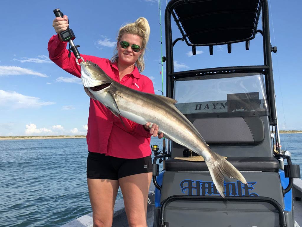 A blonde-haired woman in a red sweater holds a large Cobia aboard a center console fishing charter on a clear day, with the console behind her on the right of the image and open water on the left