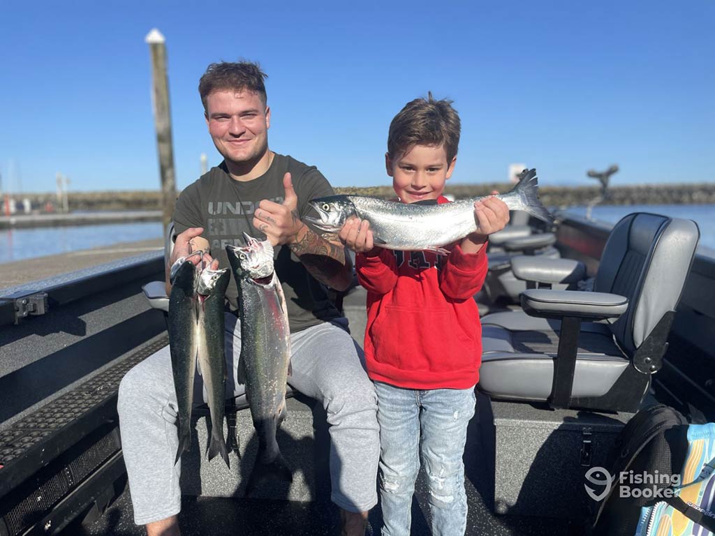 A teenager and young boy standing and holding a number of Coho Salmon on a fishing charter in Puget Sound on a clear day, with seats to the right of them