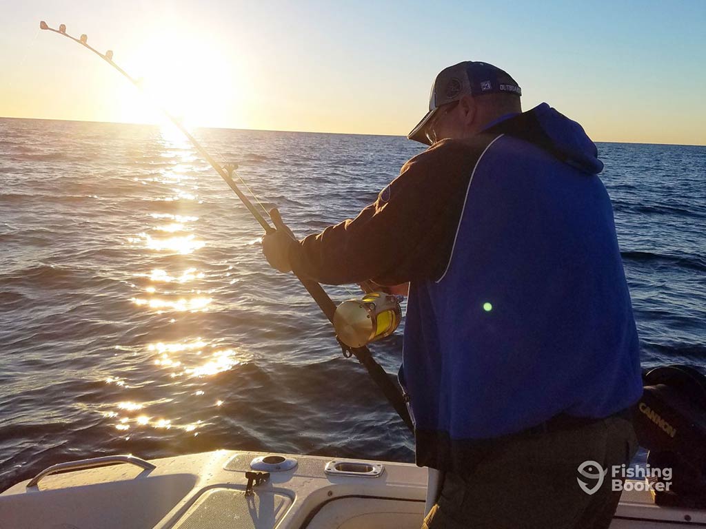 An angler struggles with an offshore fishing rod and large reel aboard a charter in the deep waters of the Gulf of Mexico on a sunny day, with the sun setting in the distance