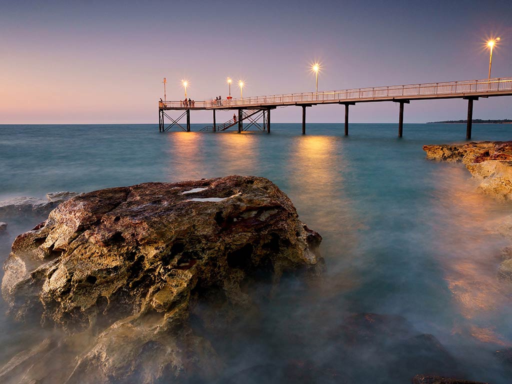 A view from the shore towards a fishing pier in Darwin, Northern Territory, at sunset, with a rock visible in the water in the foreground and the pier under lights in the distance