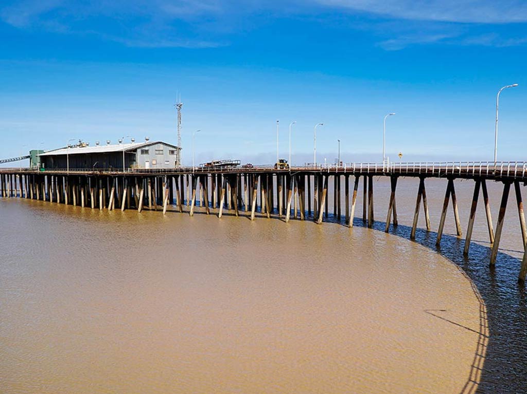 A view across muddy waters towards a big, long, wooden fishing pier in Derby, Western Australia on a sunny day