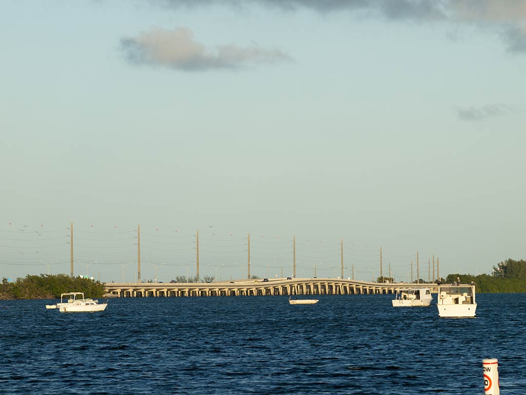 A view across the blue water towards a bridge connecting Stock Island, FL, with the rest of the Florida Keys on a sunny day, with traffic visible on the bridge and a few clouds above