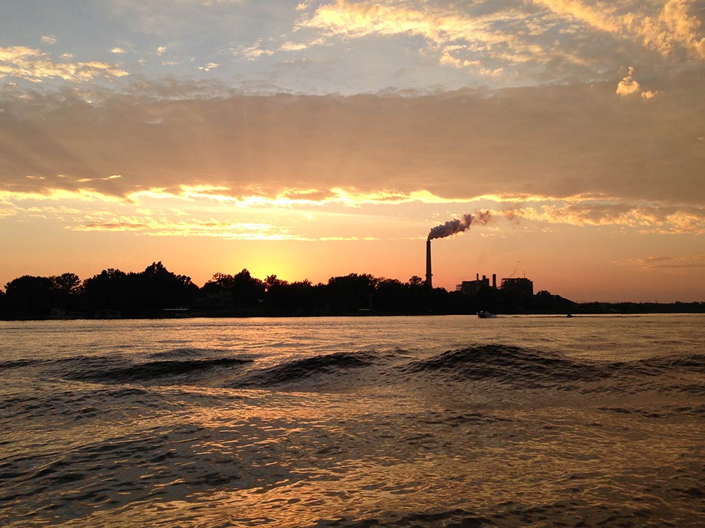 A view across the water of Lake of Egypt, Illinois, towards a power plant at sunset, with the sun setting in the distance, creating a bright orange hue and a silhouette of the trees on the shoreline and the power plant and its smoke