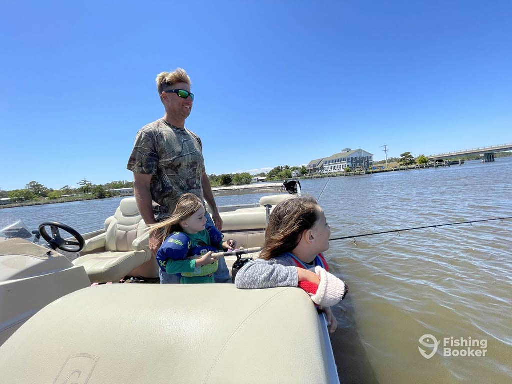 A father stands on a fishing boat, as one of his daughters fishes off the side of it in the shallow waters of Wanchese, and the other daughter looks on, perching over the side of the boat on a clear day