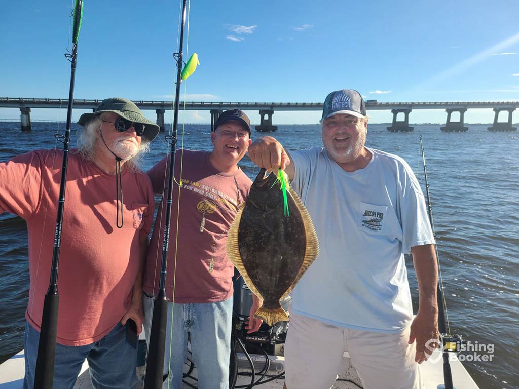 Two older men and one teenager pose on a fishing charter in front of a bridge in Wanchese, proudly presenting a Flounder they caught to the camera on a clear day