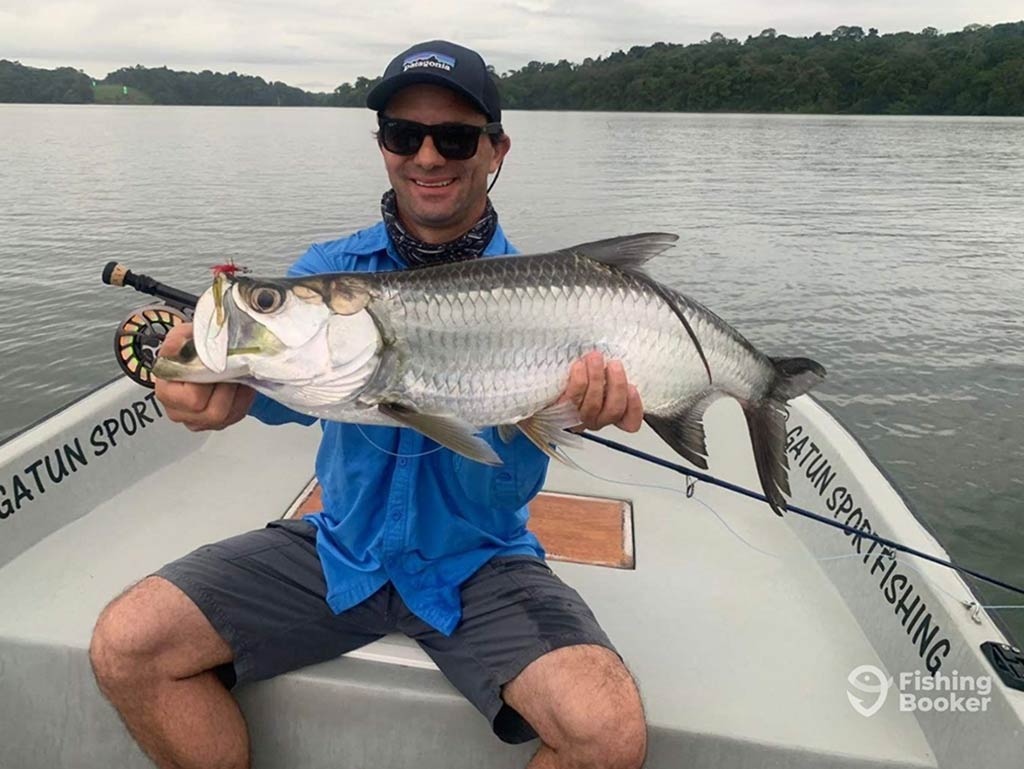 An angler sits on the front of a fishing boat, holding a Tarpon and a fly fishing rod, with the inshore waters behind him on a cloudy day