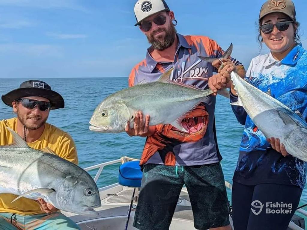 Three anglers on a fishing charter in Australia, each holding up a Giant Trevally, caught while offshore fishing on a sunny day, with the water visible behind them