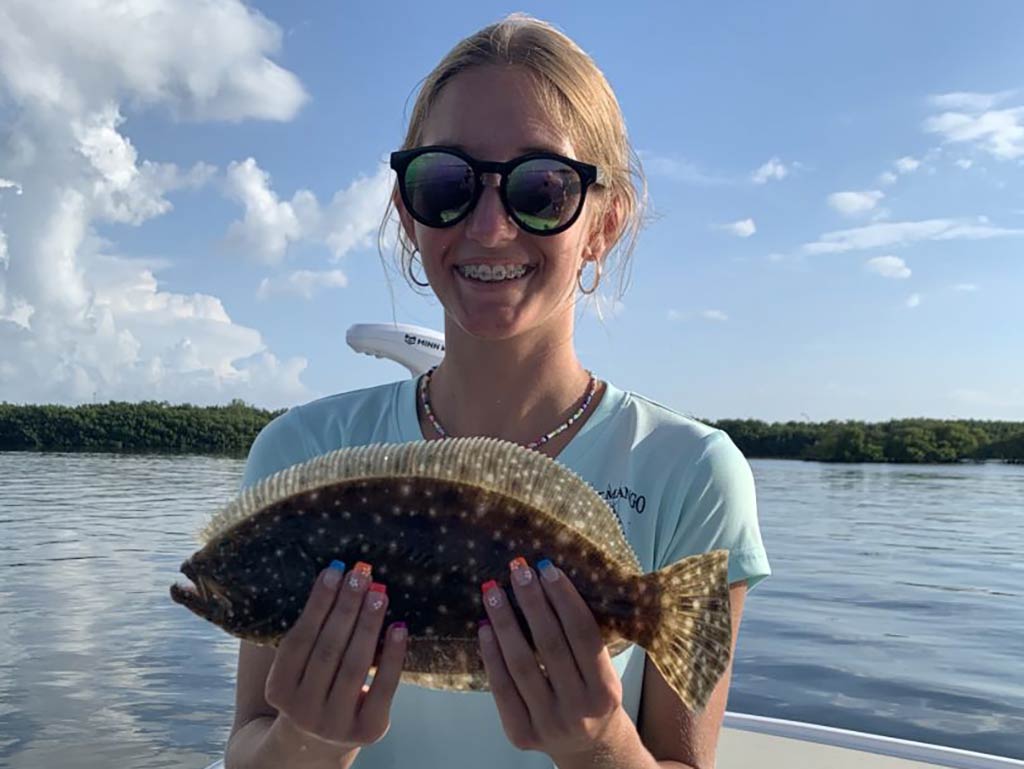 A young woman holds up her Flounder to the camera somewhere in Florida, while on a fishing trip in the calm inshore waters on a clear day