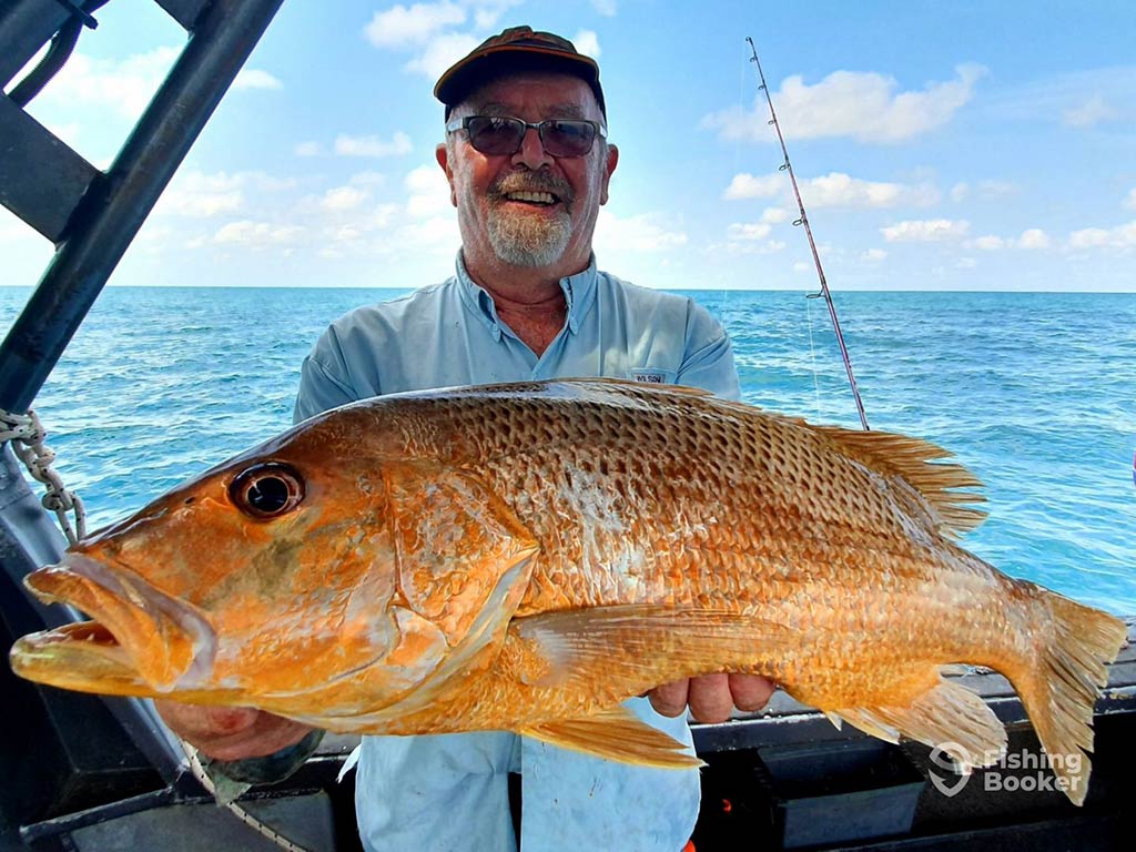 A happy angler holds a large Golden Snapper aboard a fishing charter in the Northern Territory ona sunny day, with the water and one fishing rod visible behind him