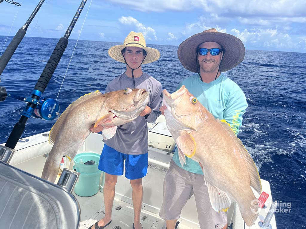 A teenage boy and a man stand on a fishing charter out of Sebastian, FL, each holding a sizeable Grouper with the water behind them on a clear day