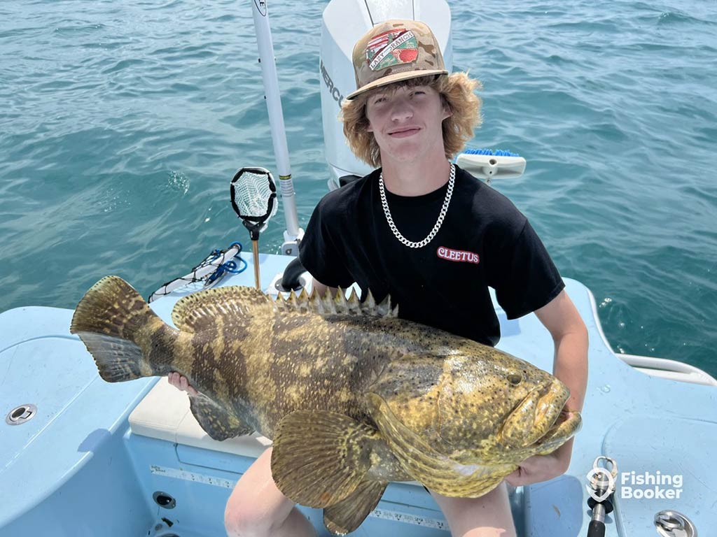 An angler with curly hair, wearing a baseball cap and sitting as he holds a Grouper caught while fishing in Stock Island on a sunny day