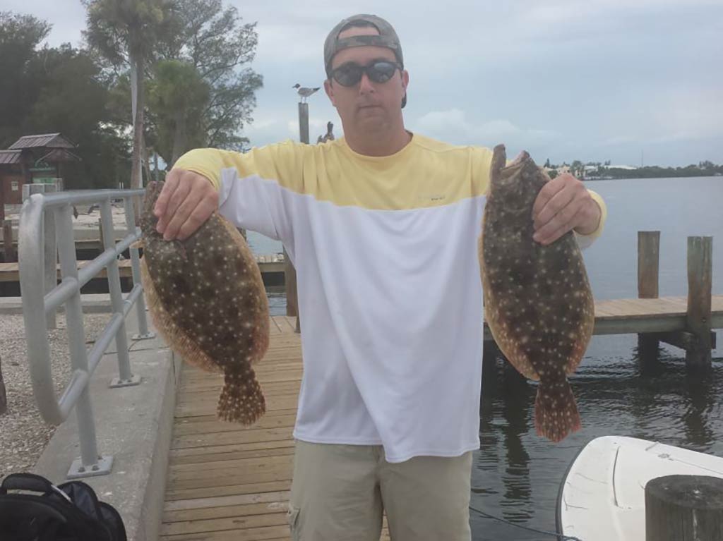 An angler in a yellow and white t-shirt holds up two Flounders while back on the dock in Florida on a cloudy day