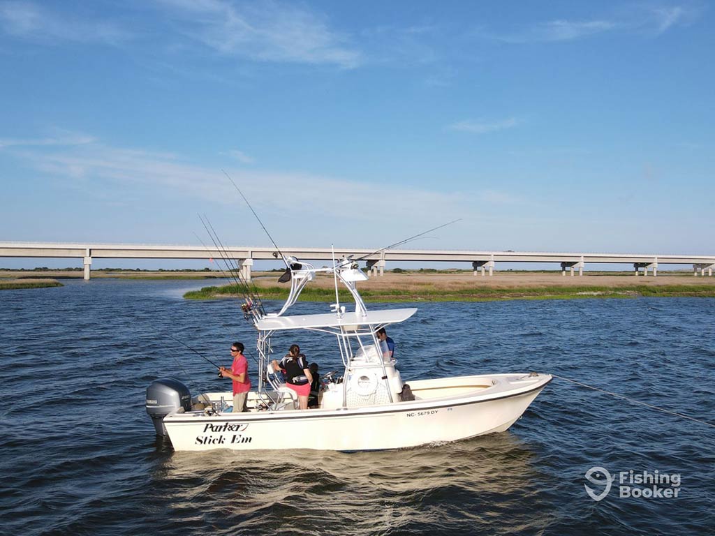 A view across the water towards a center console fishing boat, drifting along the inshore waters of Wanchese, as an angler fishes off the back of it, with a bridge and sandbar visible in the distance