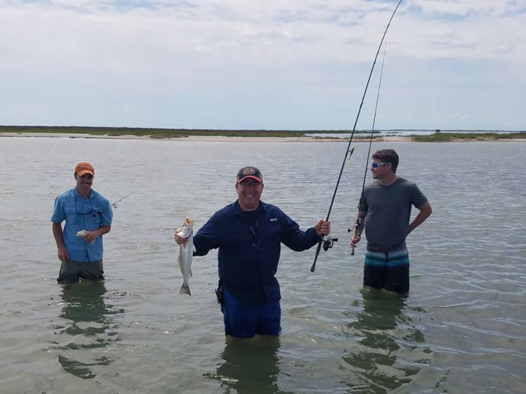 Three anglers wading in the shallow bay waters near Aransas Pass on a cloudy day, with one man in the middle holding a rod and a fish in each hand