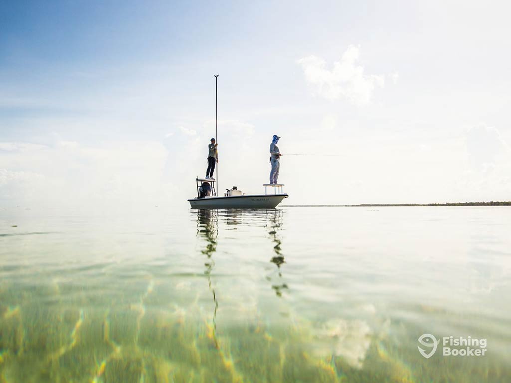 A view across the calm waters of the Florida Keys flats towards a lone fishing boat on a clear day, with a guide polling and an angler fly fishing