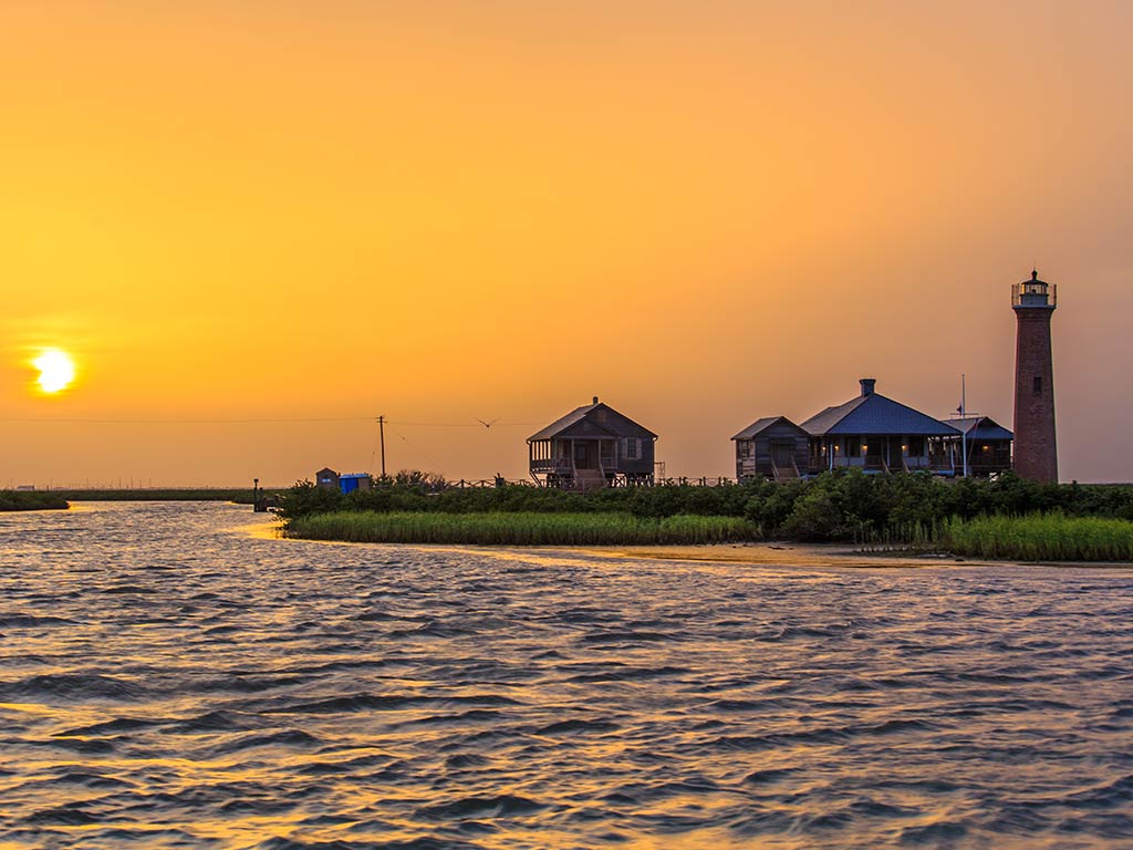 A view across the water towards a lighthouse in Port Aransas with the sun setting in the distance over the Gulf of Mexico