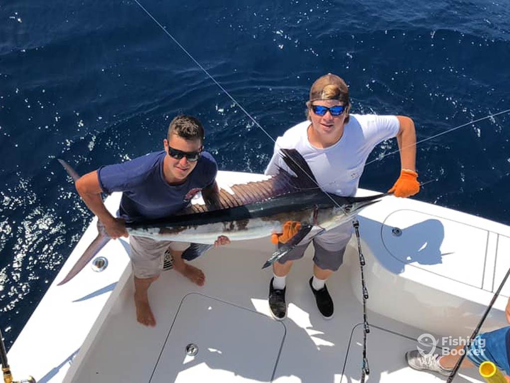 Two anglers on the deck of a fishing boat from Manteo, holding a Marlin while looking up to the camera on the flybridge, with the water behind them on a clear day