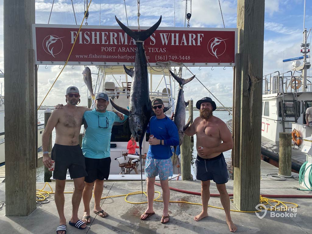 A group of anglers pose next to a record Marlin, hanging between them from a wooden board on a deck in Port Aransas, TX on a clear day