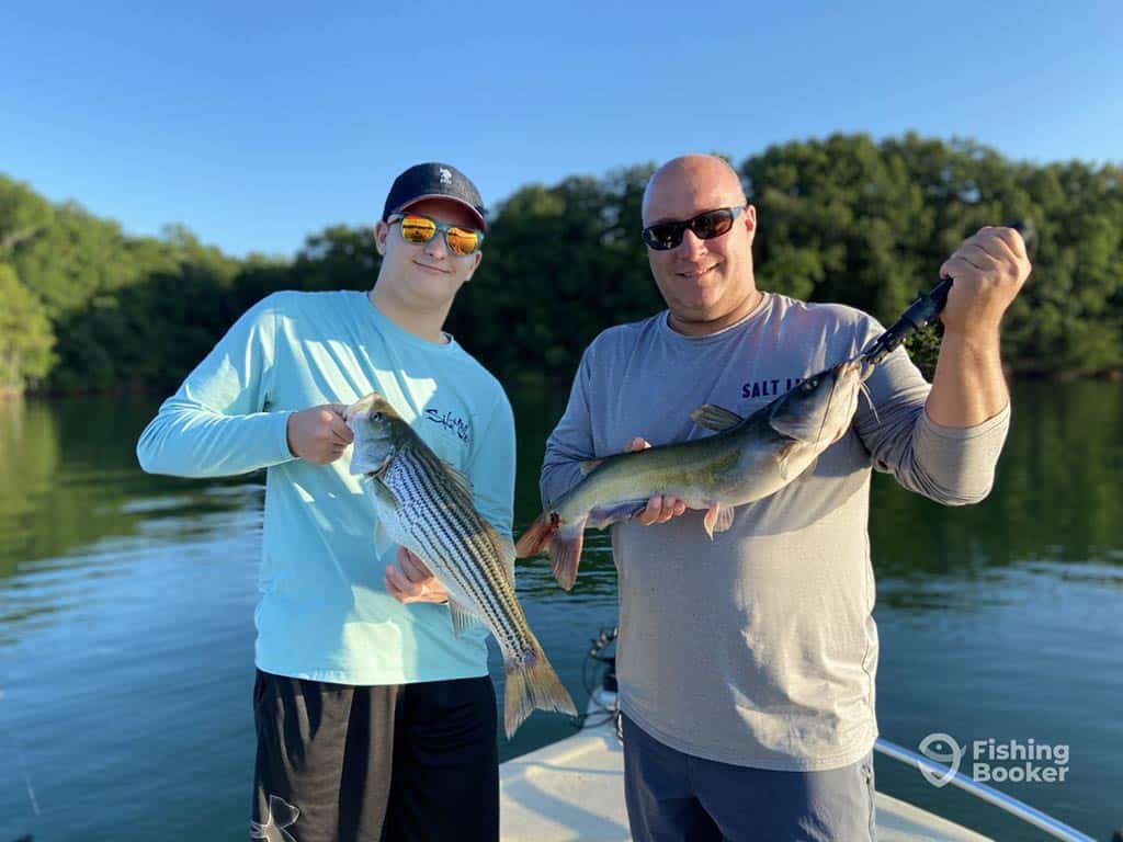 A teenager and middle-aged man stand aboard a fishing charter in Lake Lanier, one holding a Striped Bass and the other holding a Catfish on a clear day with the water and shoreline visible behind them