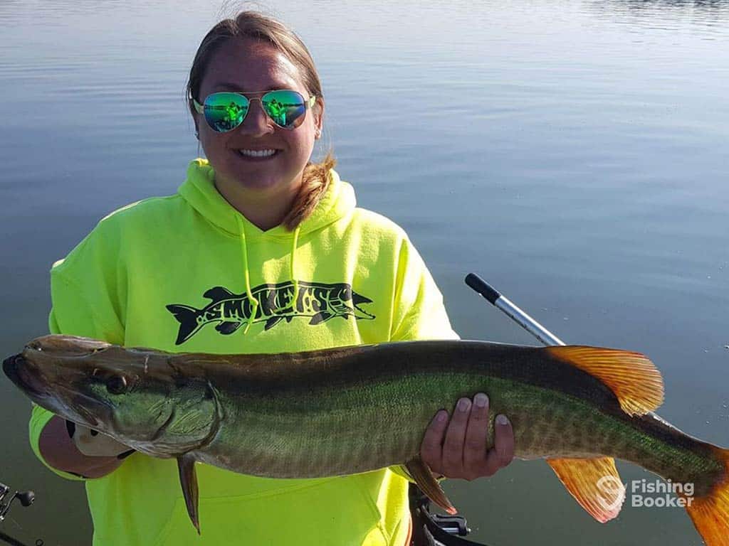 A woman in a bright lime green hoodie and sunglasses holding a Musky caught in Illinois with calm waters visible behind her