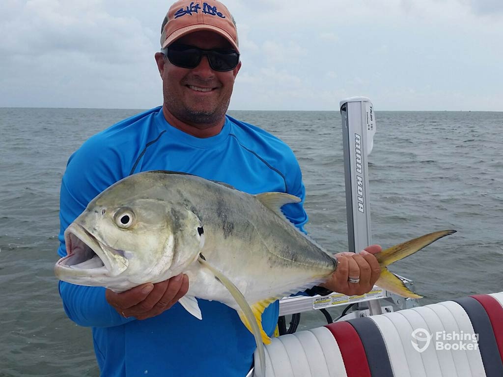 An angler in an orange baseball cap and blue shirt holds a large Jack Crevalle aboard a fishing charter out of Aransas Pass on a cloudy day, with the water behind him