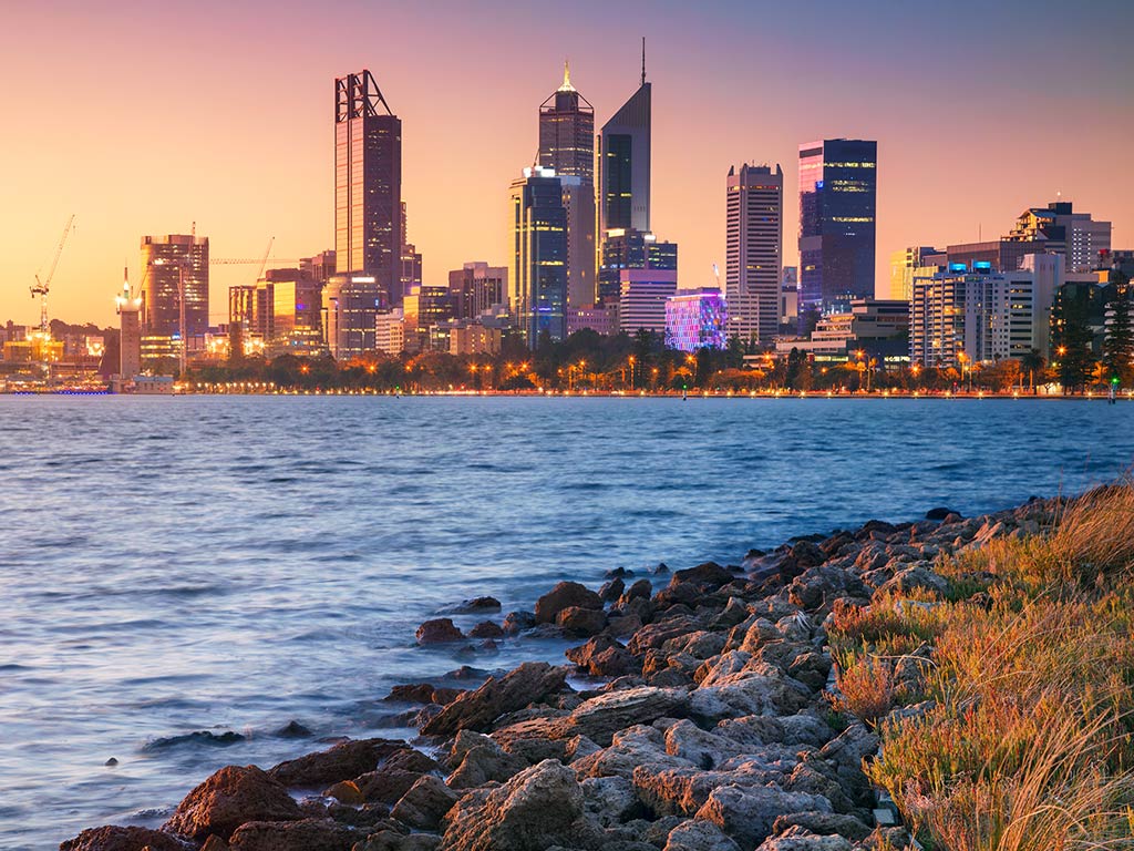 A view across the water towards the cityscape of Perth, Western Australia, at dusk, with the sky in numerous colours from purple to red