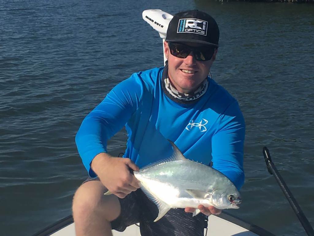 An angler in a blue shirt, crouching on an inshore fishing boat in Florida while holding a Pompano on a sunny day 