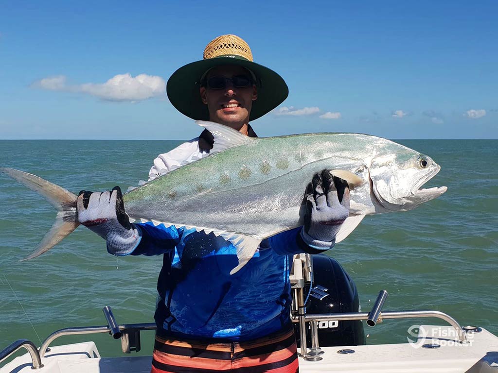 An angler in a large hat and wearing gloves holds up a large Queenfish to the camera while standing on a fishing charter in the Northern Territory on a sunny day, with the open ocean water behind him