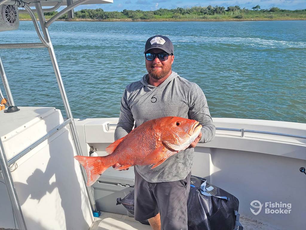 A man aboard a fishing charter near Aransas Pass, holding a Red Snapper, with the shallow waters of the bay behind him and some greenery visible in the distance on a clear day