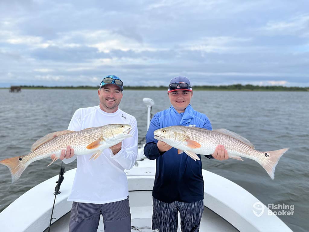 Two anglers standing side-by-side on an inshore fishing charter in Manteo, each holding a large Redfish, with the water behind them and some land visible in the distance on a cloudy day