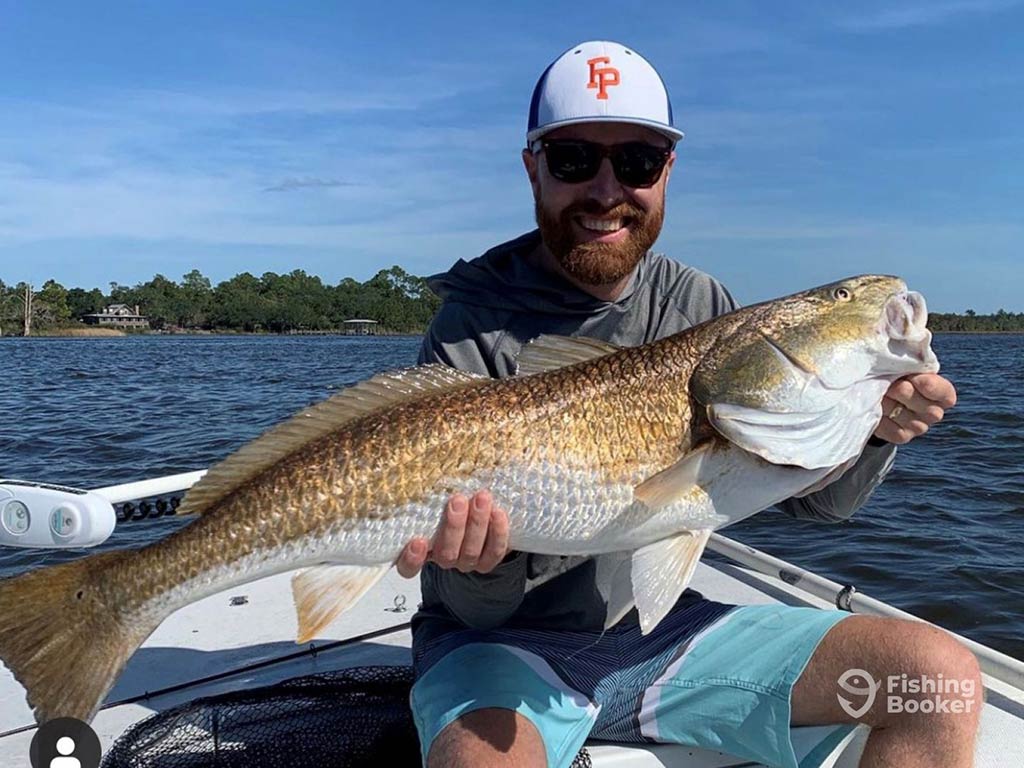 A relatively closeup shot of an angler sitting aboard an inshore charter in the Florida Panhandle and holding a Bull Redfish on a clear day with the water behind him