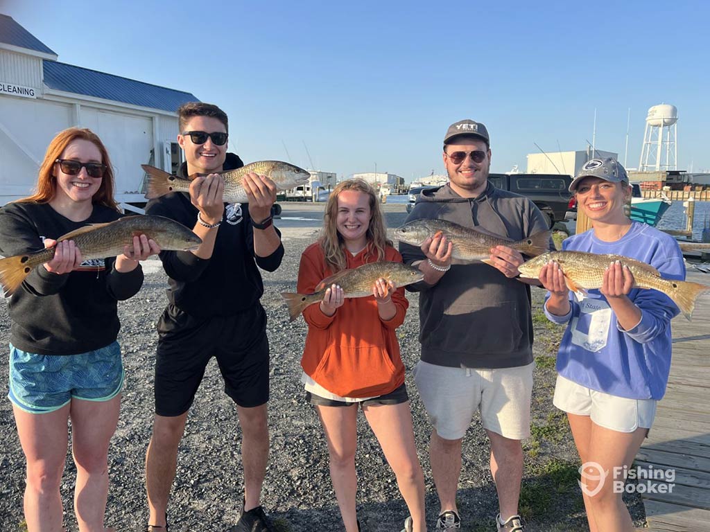 A group of anglers, probably in their early twenties, holding a Redfish each, while standing on some gravel after a successful fishing trip in Wanchese, as the sun sets on a sunny day