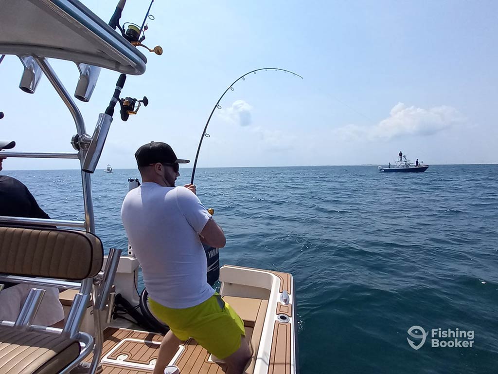 A view from behind as an angler struggles with his bent fishing rod in the nearshore waters of North Carolina with another boat visible in the distance on a clear day