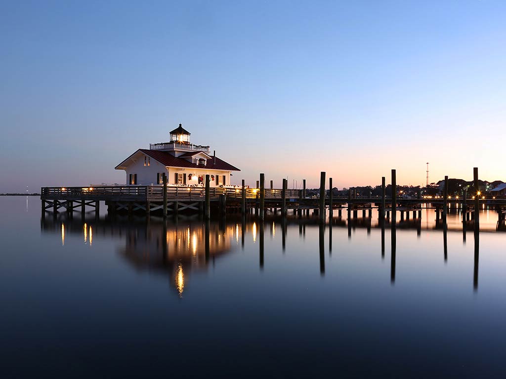 A view across the water towards Roanoke Island Lighthouse at the end of a fishing pier in Manteo, NC, at night, with a distant light from the sun visible